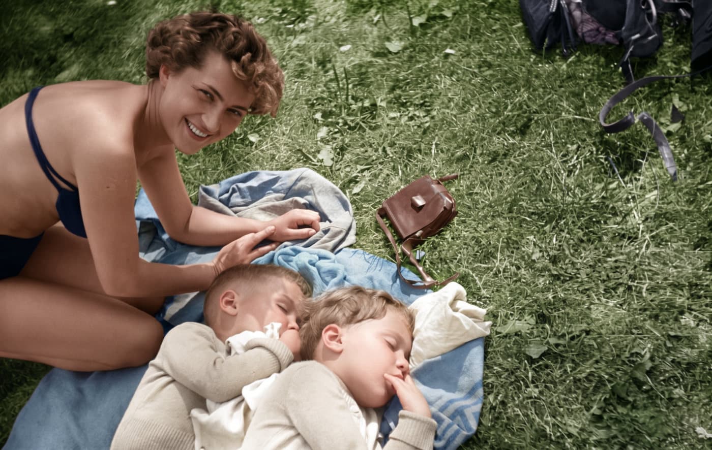 A scratched 1954 black-and-white photo of a smiling woman beside two small children asleep on a blanket in the grass, restored and colorized