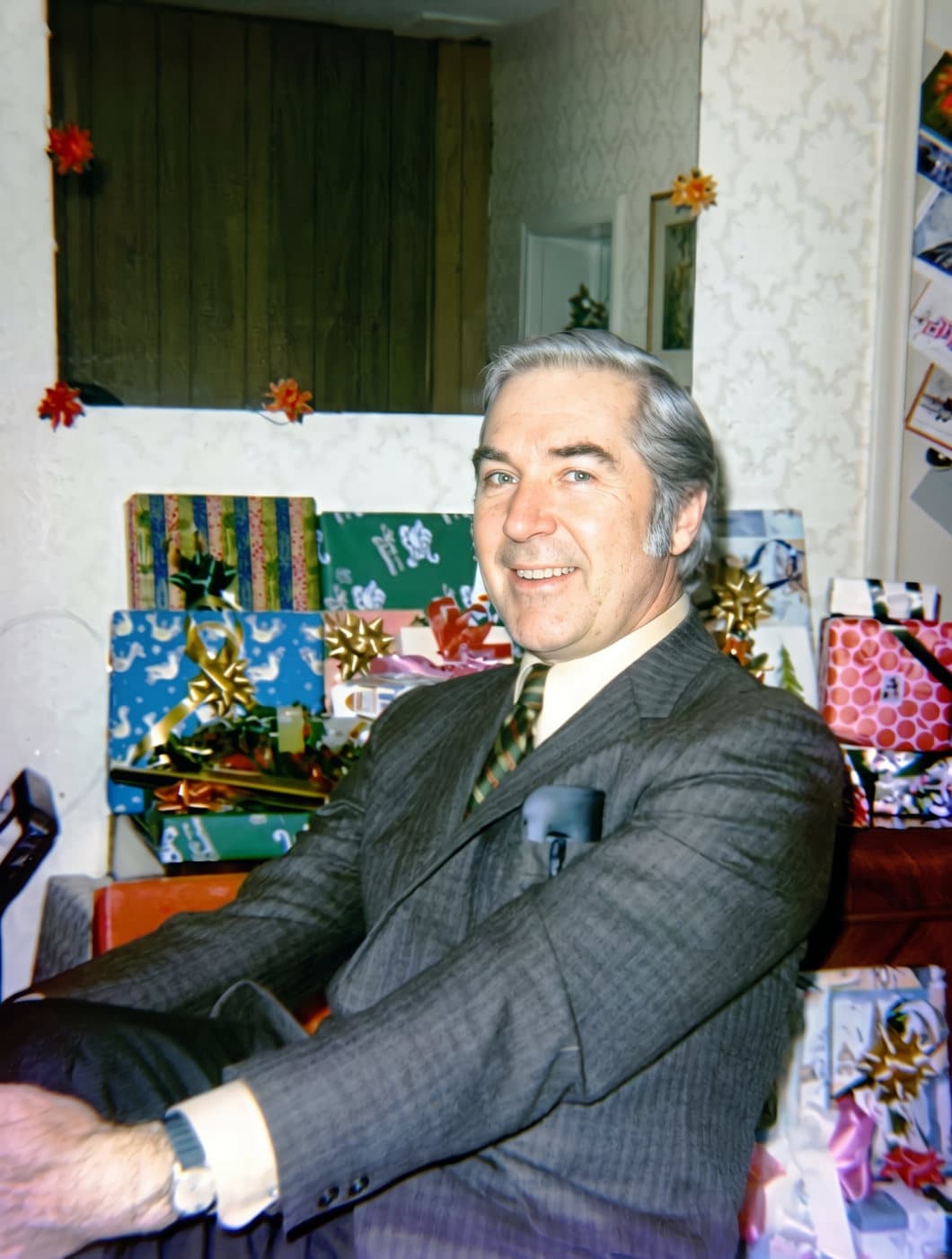 A faded 1971 Christmas snapshot of a smiling man in a suit in front of wrapped presents, with a purple colour cast, restored