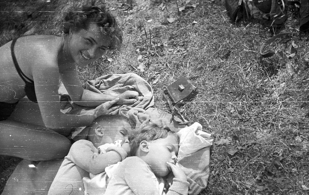 A scratched 1954 black-and-white photo of a smiling woman beside two small children asleep on a blanket in the grass, before restoration