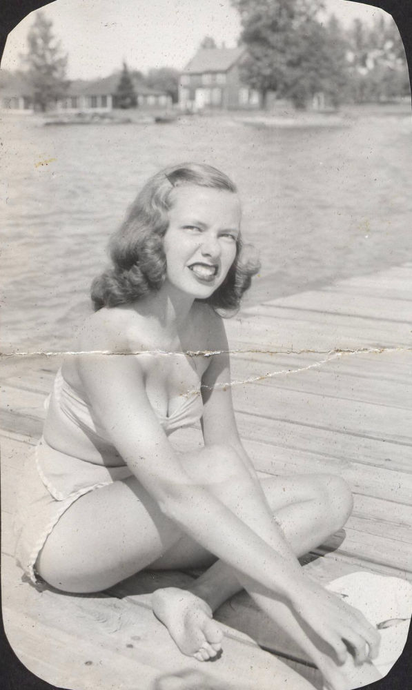 A torn 1945 black-and-white snapshot of a young woman on a lake dock, before restoration