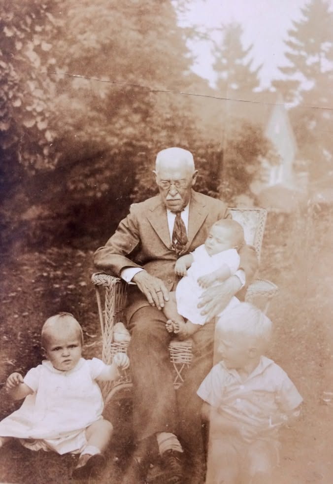 A creased sepia photo of an elderly man in a wicker chair with three small children, before upscaling