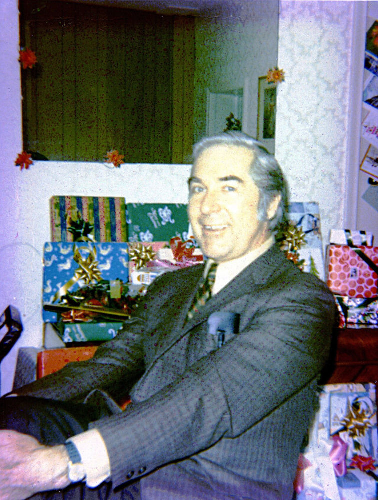 A faded 1971 Christmas snapshot of a smiling man in a suit in front of wrapped presents, with a purple colour cast, before restoration
